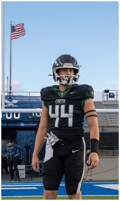 A person in a black football uniform stands on a blue field near a goalpost with a scoreboard and empty bleachers in the background. Description generated by AI