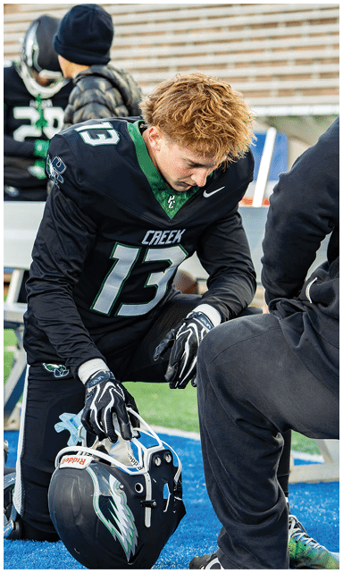 A person wearing a black football uniform with the number 13 is kneeling on a blue track while holding a football helmet. Description generated by AI