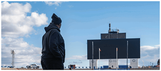 A person wearing a dark hoodie and beanie stands outdoors looking at a large blank digital billboard against a partly cloudy sky. Description generated by AI