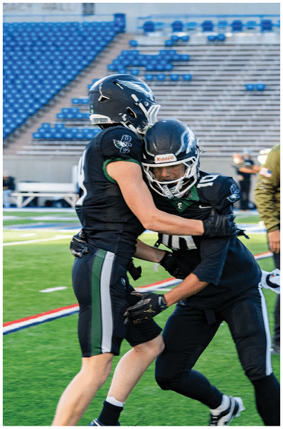 Two people wearing black football uniforms and helmets are engaged in a tackle on a football field with empty blue stadium seats in the background. Description generated by AI