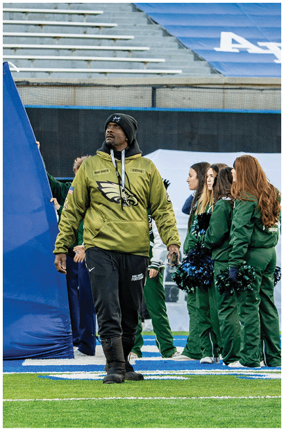 A person wearing a green Eagles jacket and black pants walks on a football field near a large blue tarp with empty stadium seats in the background. Description generated by AI