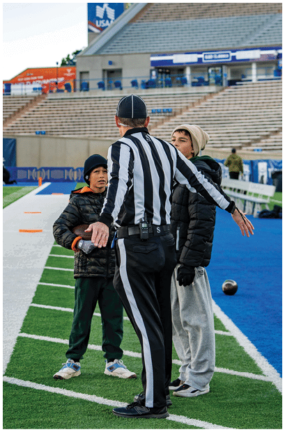 A person wearing a dark jacket, black pants, and a beanie is walking on the edge of a football field near the sideline with empty bleachers and a group of people sitting in the background. Description generated by AI