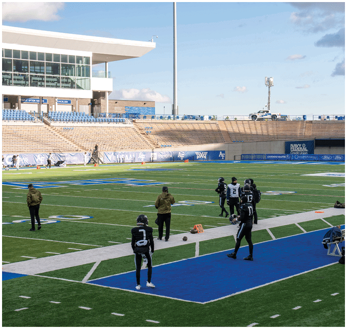 People in sports uniforms and jackets are standing and walking on a football field near the sidelines with empty stadium seats and a building in the background. Description generated by AI