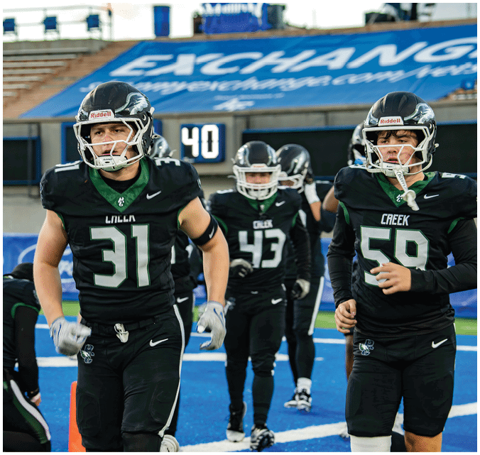 Several people in black football uniforms and helmets are on a blue field with stadium seating in the background. Description generated by AI