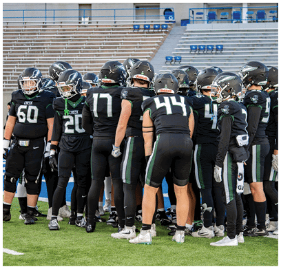A group of people wearing black football uniforms and helmets are gathered closely together on a football field with empty bleachers in the background. Description generated by AI