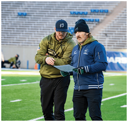 Two people wearing sports apparel and hats are standing on a football field while looking at a sheet of paper. Description generated by AI