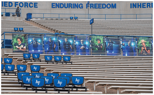 A row of large posters featuring people in sports uniforms is displayed on empty stadium bleachers with blue seats marked “AF" and a banner above reading "ENDURING FREEDOM." Description generated by AI