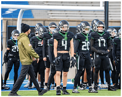 A group of people in black football uniforms and helmets stand on a field near a goalpost while one person in a green jacket and black beanie walks past them. Description generated by AI