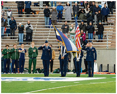 Adults in casual and military-style clothing stand in bleachers and on the ground with some saluting and others holding flags, suggesting a patriotic or ceremonial event. Description generated by AI