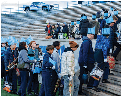 People wearing blue band uniforms and hats are gathered on stadium steps, some holding musical instruments, while others sit in the bleachers. Description generated by AI