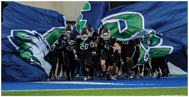 People wearing helmets are gathered in front of large blue and green flags waving in the background. Description generated by AI