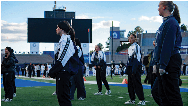People wearing athletic clothing stand on a sports field with a large scoreboard and stadium seating in the background under a partly cloudy sky. Description generated by AI