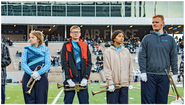 Four people wearing band uniforms and gloves stand on a field holding brass instruments with a stadium crowd in the background. Description generated by AI