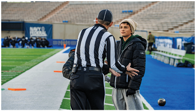 A person wearing a striped referee uniform is gesturing while talking to a person in a beanie and puffy jacket standing on the sidelines of a sports field. Description generated by AI
