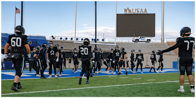 A group of people wearing black football uniforms with helmets are gathered on a blue and green football field near the stands. Description generated by AI