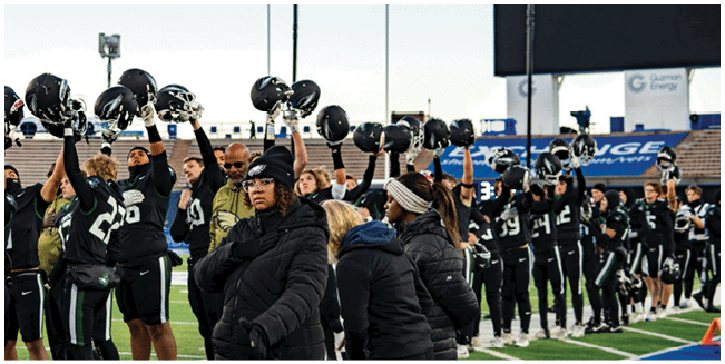 People in sports uniforms stand on a field holding helmets raised above their heads while others in warm clothing stand nearby. Description generated by AI