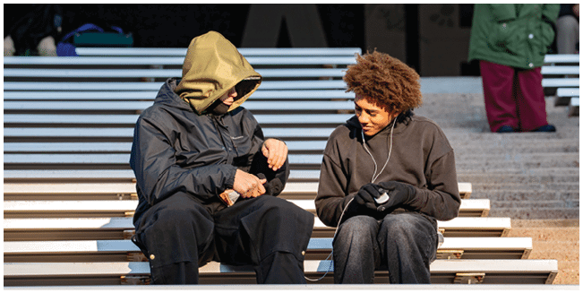 Two people dressed in warm clothing are sitting on outdoor bleachers, engaged in conversation with one person wearing earphones. Description generated by AI