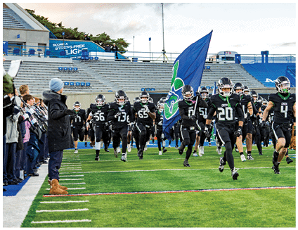A group of football players in black uniforms with helmets run onto a field while one player carries a large blue flag with a green and white emblem, and people stand on the sidelines watching. Description generated by AI