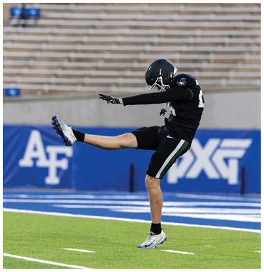A person in black football gear is kicking a ball on a field with empty bleachers in the background. Description generated by AI