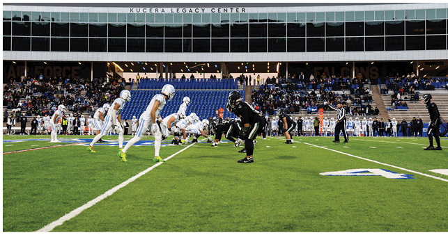 Two teams of people in sports uniforms are lined up facing each other on a football field inside a stadium with spectators in the stands. Description generated by AI