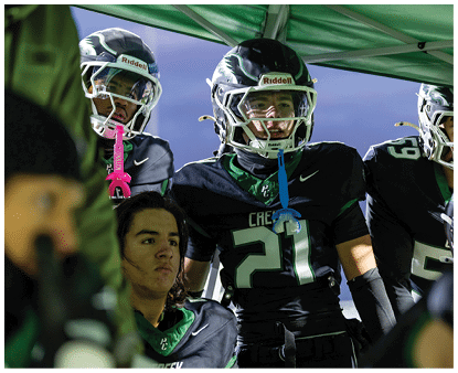 Two people wearing football helmets and uniforms stand under a green canopy while another person with long hair looks on nearby. Description generated by AI