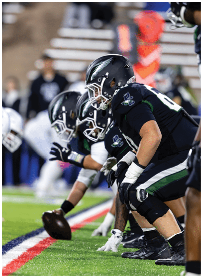 Two opposing American football teams in helmets and uniforms are lined up facing each other on a field, ready for the snap of the ball. Description generated by AI