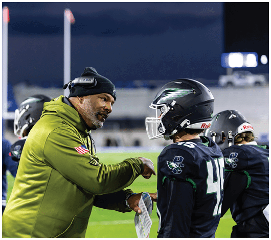 A person wearing a green hoodie and headset is talking to a football player in a black helmet and jersey on a field at night. Description generated by AI