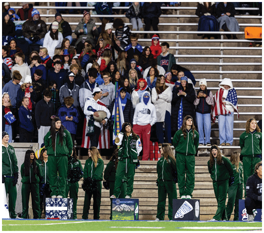 People in green athletic uniforms stand on a field in front of a crowd of spectators seated on bleachers during an outdoor event. Description generated by AI