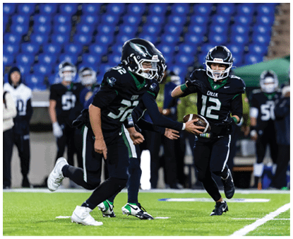 Two people in black football uniforms are on a field with one handing the ball to the other while several others stand in the background near blue stadium seats. Description generated by AI