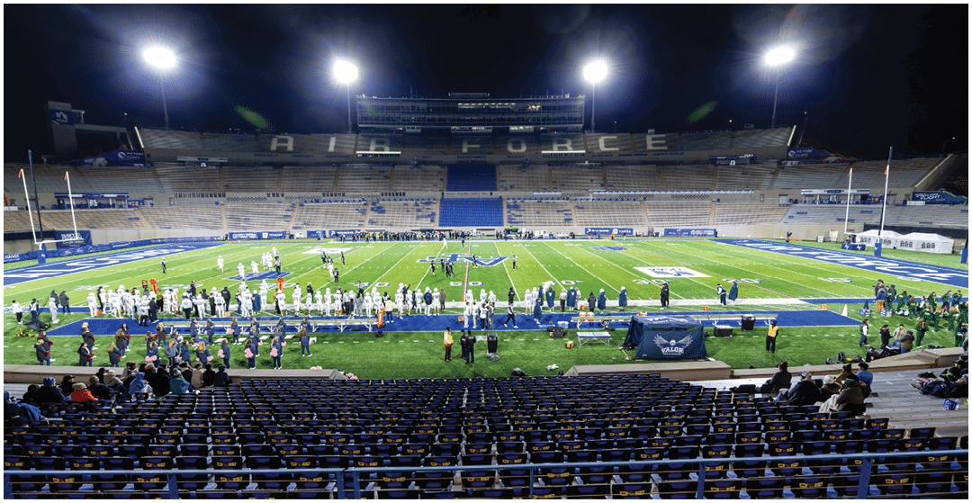 A nighttime football game is taking place in a large stadium with players on the field, people on the sidelines, and spectators seated in the stands. Description generated by AI
