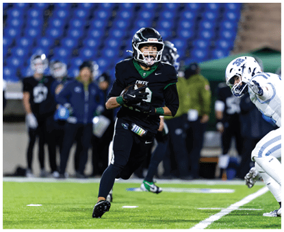 A person in black sports gear and helmet is running on a football field holding a football while another person in white sports gear is approaching from the right. Description generated by AI