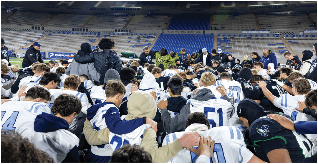 A large group of people in sports uniforms are gathered closely together in a stadium, appearing to be in a moment of team huddle or prayer. Description generated by AI