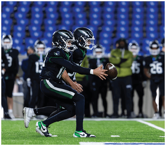 Two people in football uniforms and helmets are on a field with one handing off a football to the other while teammates and a coach stand in the background. Description generated by AI