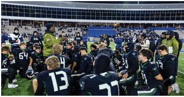 A group of people wearing football uniforms are gathered on a field listening to a person in a green jacket giving instructions during a game or practice. Description generated by AI