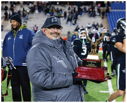 A person wearing a gray jacket and black cap is smiling and holding a trophy on a sports field with other people and players in the background. Description generated by AI