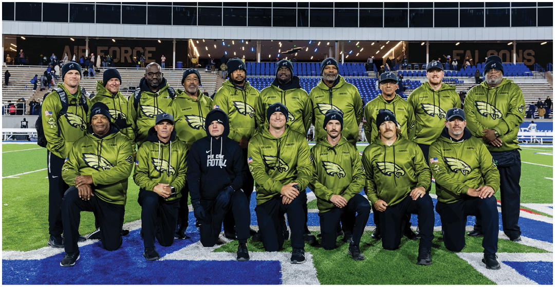 A group of people wearing matching green jackets and black pants pose together on a football field in front of stadium seating under a sign that reads Kucera Legacy Center. Description generated by AI