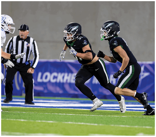 Two people in black football uniforms are running toward a person in a white football uniform on a field with a referee in the background. Description generated by AI
