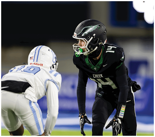 Two people in football uniforms and helmets are facing each other on a field, appearing ready for a play. Description generated by AI