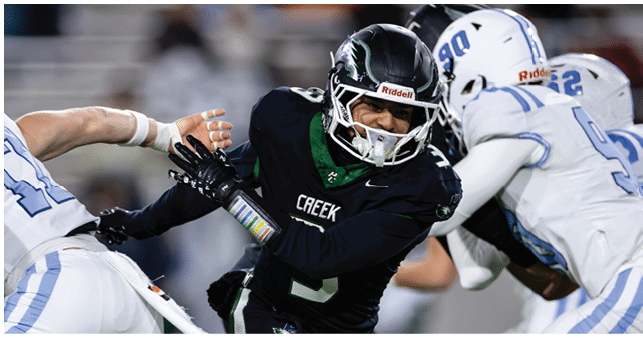 A person in a black football uniform with “CREEK" on the chest is running while two people in white football uniforms attempt to block or tackle them during a game. Description generated by AI
