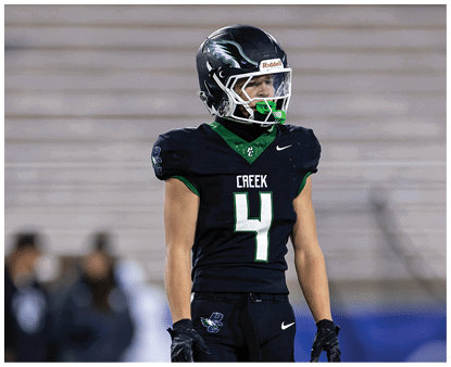 A person wearing a black football uniform with the number 4 and the word Creek stands on a field with blurred spectators in the background. Description generated by AI