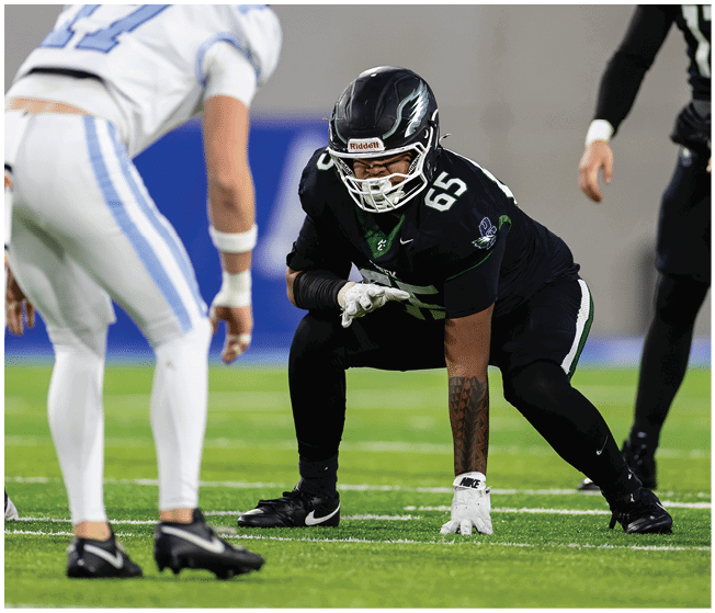 Two people in football uniforms are positioned on a field facing each other, with one in a black uniform crouched in a ready stance and the other in a white uniform standing slightly bent forward. Description generated by AI
