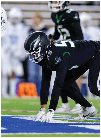 A person in a dark football uniform and helmet is crouched in a ready position on a blue and white field with other players blurred in the background. Description generated by AI