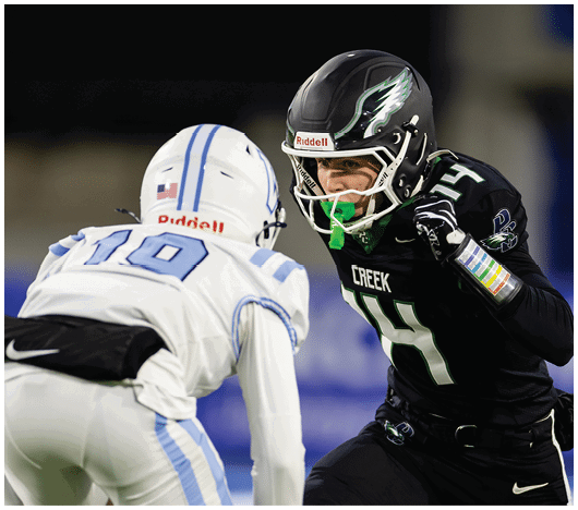 Two football players in helmets and uniforms face each other on the field during a game, one wearing black with “CREEK" on the jersey and the other in white with blue stripes. Description generated by AI