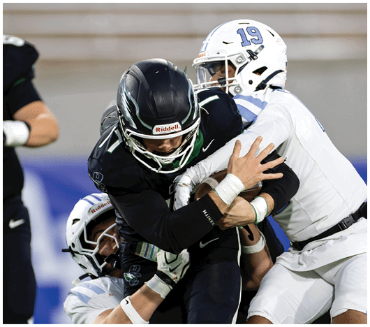 Three people in football uniforms are engaged in a tackle during a game, with one person in black being held by two people in white. Description generated by AI