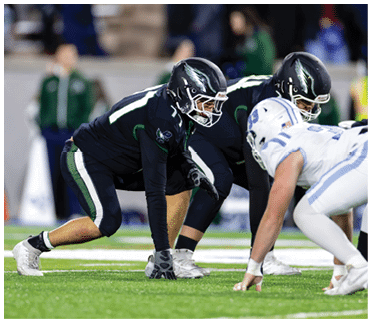 Two football players in black uniforms are crouched on the field facing two players in white uniforms in a game setting. Description generated by AI
