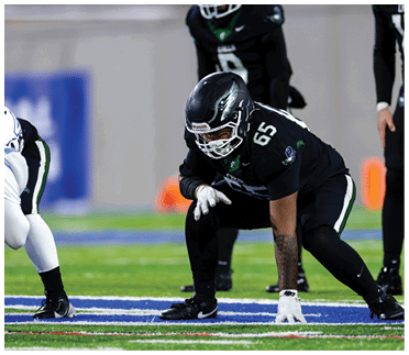 A person in black football gear with the number 65 is crouched in a ready position on a football field facing an opponent in white gear. Description generated by AI