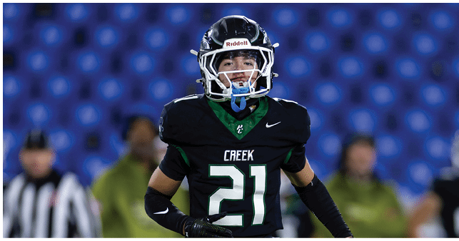 A person wearing a black football uniform with the number 21 and the word Creek on the chest is standing on a field with blurred people and blue stadium seats in the background. Description generated by AI