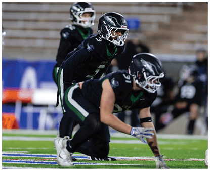 Two people in black football uniforms and helmets are positioned on a field, with one crouching and the other standing behind, preparing for a play. Description generated by AI