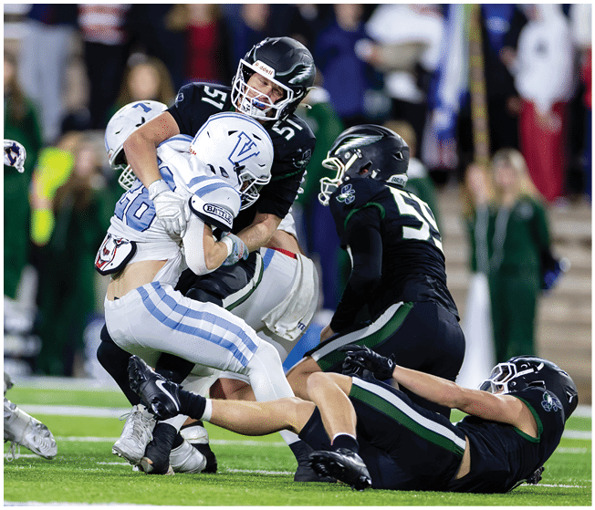 Three football players in black uniforms tackle a player in a white and blue uniform on a green field during a game. Description generated by AI