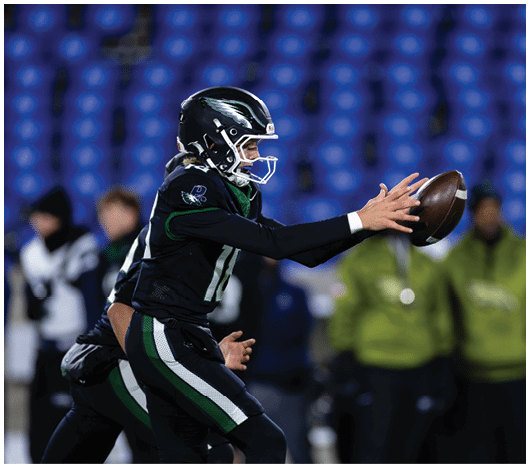A person wearing a black football uniform and helmet is reaching out to catch a football on a field with blurred spectators in the background. Description generated by AI